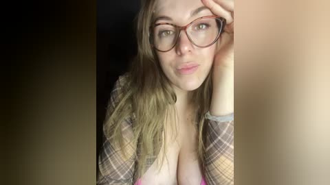 Video of a young woman with light skin, green eyes, and long brown hair, wearing red-framed glasses and a plaid shirt, partially unbuttoned, revealing cleavage.
