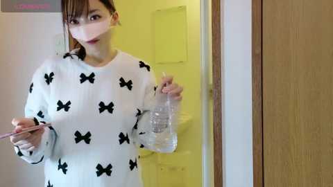 A video of an Asian woman with light skin and straight brown hair, wearing a white sweater with black bow patterns and a mask, holding a phone and a plastic bottle, standing in a room with a yellow wall and a wooden door.