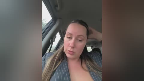 Video of a fair-skinned woman with long, blonde hair in a pinstriped shirt, sitting in a car, looking slightly annoyed, with blurred background.