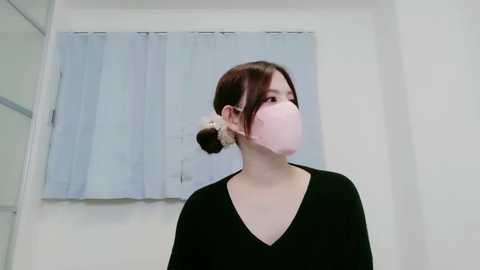 A video of a young Asian woman with pale skin, dark hair tied in a low bun, wearing a pink face mask and black V-neck sweater, standing in a white-walled room with light blue curtains.
