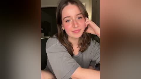 Video of a young woman with fair skin and brown hair, wearing a gray t-shirt, leaning on her elbow, smiling softly. Background features a blurred indoor setting with neutral tones.