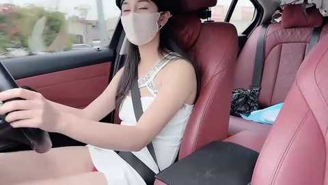 Video of an Asian woman with long black hair, wearing a white dress, mask, and driving a car with red leather seats.