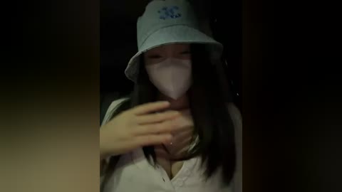 A video of a young Asian woman with long black hair, wearing a light blue bucket hat and white face mask, standing indoors with a dimly lit background.