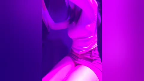 Video of a woman with fair skin and dark hair, wearing a tight, shiny pink top and shorts, illuminated in vibrant pink and purple light. Her arms are raised, body slightly twisted.