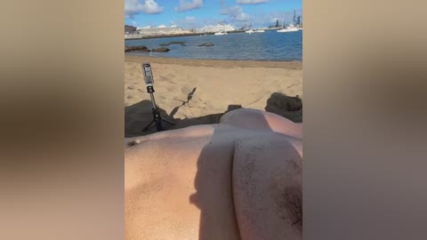 A video of a sandy beach with blue water, rocky outcrops, and distant boats under a clear sky. The image is taken from a person lying face down, with their buttocks and lower back visible.
