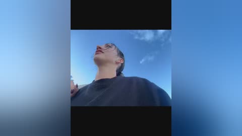 Video of a young woman with short brown hair, wearing glasses, looking upward at the sky. She has a fair complexion and is dressed in a dark shirt. The background features a clear blue sky with a few clouds.
