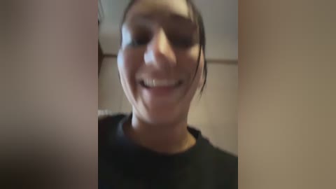 A blurry video of a smiling young woman with dark hair and light skin, wearing a black top, captured indoors.