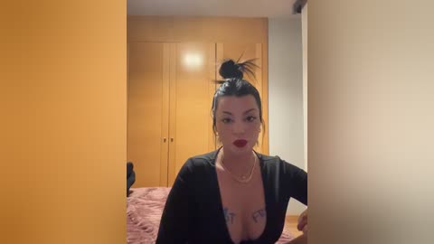 Video of a woman with dark hair styled in a messy bun, wearing a black dress, sitting on a bed. Background shows a wooden wardrobe and beige walls.
