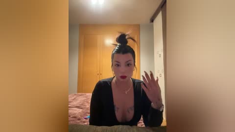 A video of a Latina woman with dark hair in a messy bun, wearing a black top with a deep V-neckline, standing in a dimly lit bedroom with beige walls and wooden doors.