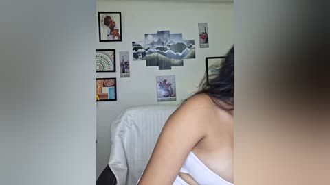 Video of a woman with medium skin tone, wearing a white bra, standing with her back to the camera, surrounded by framed videos on a white wall.