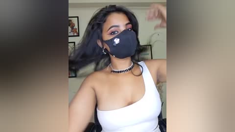 A video of a young woman with medium brown skin and long black hair, wearing a black face mask, white one-shoulder top, and black choker. She's indoors, with a blurred background showing framed art and a plant.