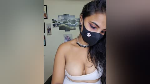 Video of a young woman with long dark hair, wearing a black face mask, white tube top revealing cleavage, and a choker necklace, in a room with framed photos and a plant poster on the wall.