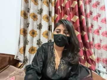 Video of a South Asian woman with medium skin tone and long black hair, wearing a black face mask, black blouse, and gold necklace, sitting in front of floral-patterned curtains.
