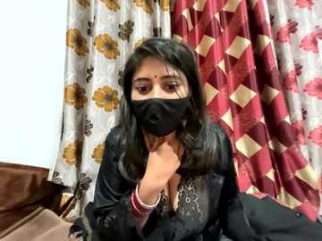 Media: Video of a South Asian woman with long black hair, wearing a black face mask, black top, and red bangles, seated indoors. Curtains with floral and diamond patterns in white, gold, and red are visible in the background.