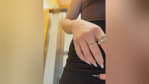 A video captures a close-up of a woman's hand and arm in a black dress, showing long, manicured nails with a silver ring and a diamond ring. The background features a beige wall and a staircase railing.