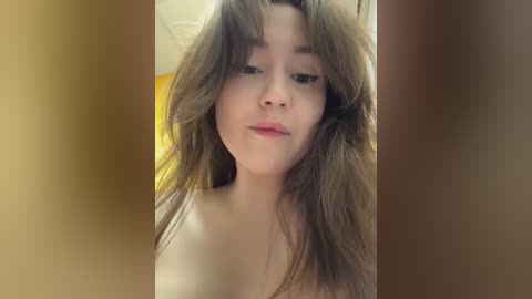 Video of a young woman with fair skin, light brown hair, and light makeup, smiling slightly. Background is blurred, suggesting an indoor setting with warm lighting.