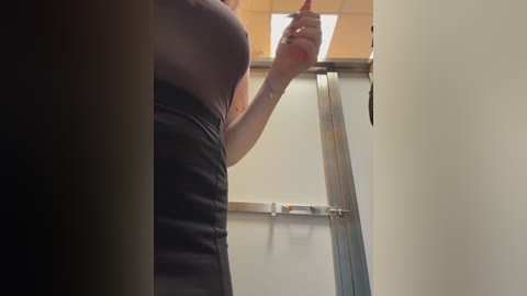 A video shows a woman with fair skin, wearing a black skirt and dark top, reaching for something on a high shelf in a brightly lit room with beige walls and a metal ladder.