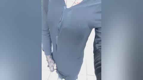 A video of a person wearing a light blue, long-sleeved button-up shirt, standing in front of a tiled wall. The image is slightly blurred, with a focus on the shirt's texture and the person's upper body.