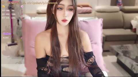 Video of a young Asian woman with long, straight brown hair, wearing a black, see-through, off-shoulder top, sitting indoors against a beige sofa. The background is blurred, featuring a modern living room.