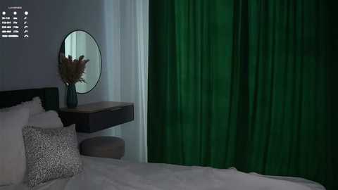Video of a modern bedroom with a dark wooden headboard, white bed, green velvet curtains, and a round mirror with a wooden frame above a dark wooden nightstand.