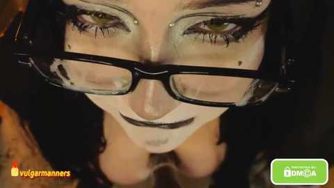 Close-up video of a woman with fair skin, wearing black-framed glasses, heavy makeup, and a black top. The background is blurred. Watermarked with \"vulgumonsters\" and \"IMAGICA.\