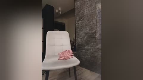A video of a minimalist room with a white modern chair, a pink crocheted blanket draped over the back, and a textured grey brick wall in the background.