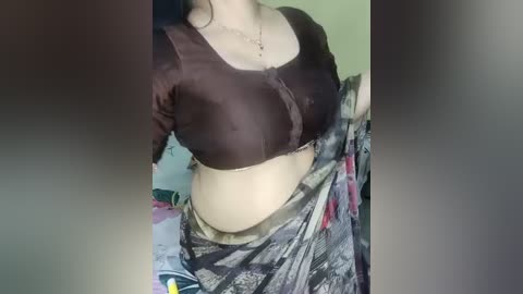 Video of a woman with fair skin, wearing a tight, dark brown crop top and a patterned skirt, partially visible. Background shows a blurred indoor setting with green walls and indistinct objects.