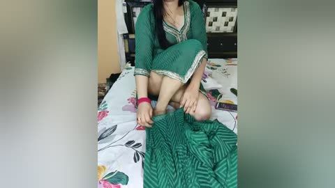 A video of a South Asian woman with medium skin tone, sitting on a bed with floral sheets. She wears a green, embroidered dress and a pink bracelet, holding a green, textured blanket. The background shows a tiled wall and bookshelves.