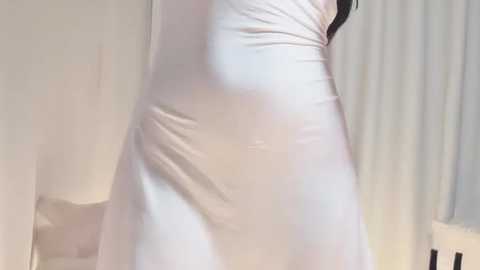 Video of a woman's lower body in a white, fitted, sleeveless dress, showing a slight curve at the waist and a hint of her legs. Background features a white wall and a white curtain.