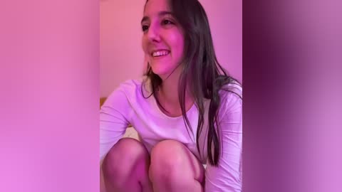A video of a young woman with long dark hair, smiling, sitting on a floor with a white shirt, in a softly lit room with pink and purple hues.