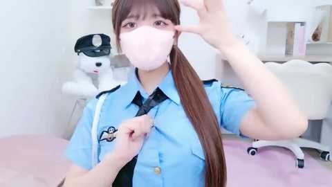 Video of a young Asian woman with long brown hair in a ponytail, wearing a blue police uniform, mask, and gloves, adjusting her mask in a brightly lit, white-walled room with a plush toy dog and white chair.