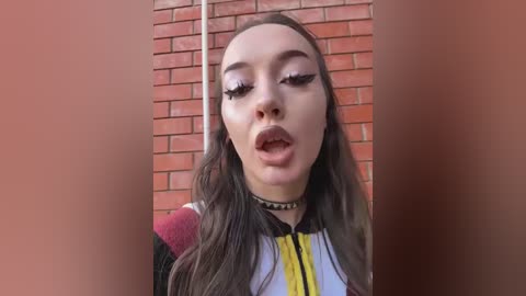 Video of a young woman with long, straight brown hair, wearing a black choker and a colorful, patterned top, against a red brick wall, looking surprised with open mouth and exaggerated facial expressions.
