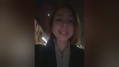 Video of a smiling young woman with shoulder-length brown hair, wearing a black coat, taken in dimly lit, blurred urban background with streetlights.