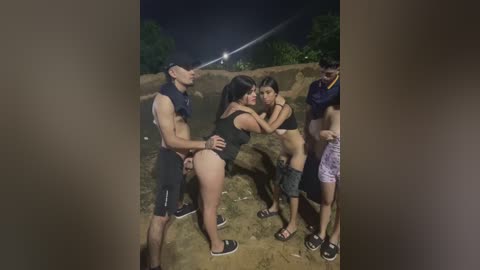 Video of a group of four shirtless men and two women in swimsuits, embracing playfully on a sandy beach at night, with dim lighting.