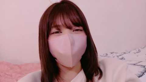 Video of a young Asian woman with straight, shoulder-length brown hair, wearing a white surgical mask covering her nose and mouth. She has light skin and is seated against a pink and white background with a subtle floral pattern.
