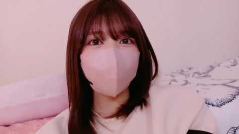 Video of an Asian woman with straight, shoulder-length brown hair, wearing a white surgical mask, sitting on a bed with white and pink linens, in a softly lit room.