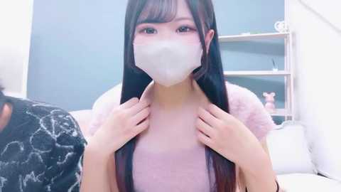 Video of an Asian woman with long, straight black hair, wearing a white face mask and light pink top, touching her collar. Background features a white room with a shelf and small decorative items.