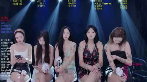Video of five Asian women in casual attire seated on stage under bright lights, each with a small white flower, displaying various expressions.
