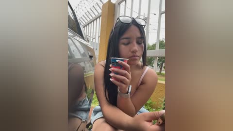 A video of a young woman with long dark hair, wearing a sleeveless top, sitting in a glass greenhouse, holding a phone.