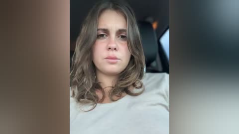 Video of a young woman with wavy brown hair, wearing a white shirt, giving a neutral expression, taken indoors with blurred background.