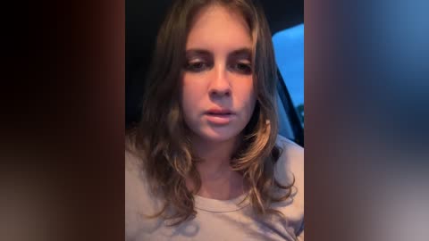 Video of a young woman with long, wavy, light brown hair, wearing a light-colored top, sitting indoors with a somber expression.