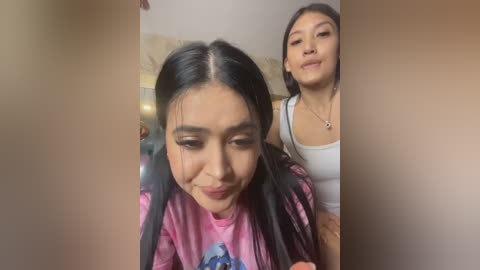 Video of two women in a cozy indoor setting: one with long black hair and a pink shirt, the other with medium-length black hair and a white top.