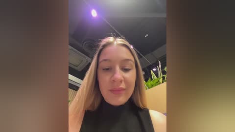 A video of a young woman with long blonde hair, wearing a black top, standing indoors with a bright spotlight above, creating a dramatic effect.