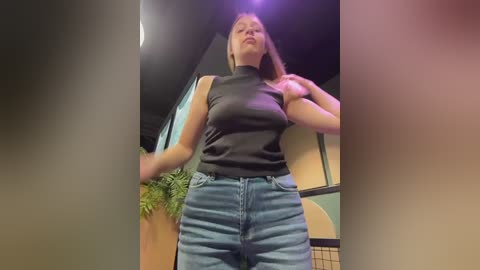 A video of a Caucasian woman with fair skin, wearing a sleeveless black turtleneck and high-waisted blue jeans, standing indoors in a modern room with a large green plant and a black ceiling.