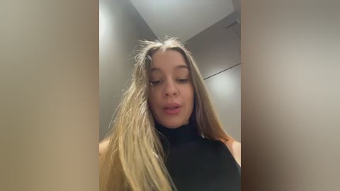 Video of a young woman with long, messy blonde hair, wearing a black sleeveless top, standing indoors with beige walls and a white ceiling.