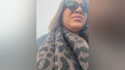 A video of a plus-sized woman with medium brown skin, shoulder-length brown hair, and dark sunglasses. She wears a leopard-print scarf over a dark coat, smiling with her lips slightly parted.