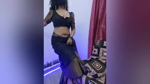 A video of a woman with medium brown skin, wearing a black lace bra and matching skirt, standing against a backdrop of a bed with patterned sheets and a red curtain.