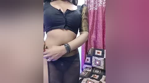 A video of a woman with medium skin tone wearing a black crop top and sheer black pants, standing in a room with a pink curtain and patterned fabric in the background.