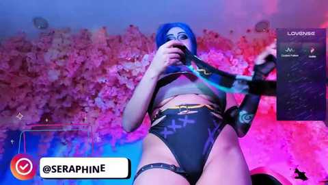 Video of a woman with blue hair in a dark, tight, cross-patterned bodysuit, holding a microphone, against a colorful, abstract background.