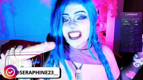 Video of a woman with vivid blue hair and dark makeup, wearing a black top, holding a large, erect penis. Background includes pink and blue lights, a television screen, and social media logo.